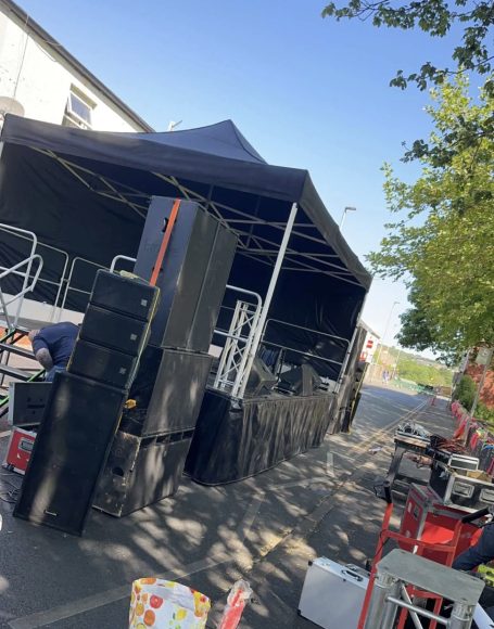 A stage setup with speakers, a black canopy, and equipment in a sunny outdoor area.